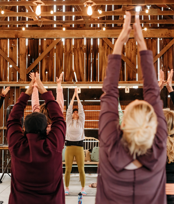 A group of people practice yoga indoors, standing with arms raised. Warm lights hang from the wooden ceiling, and sunlight filters through wooden slats on the walls.