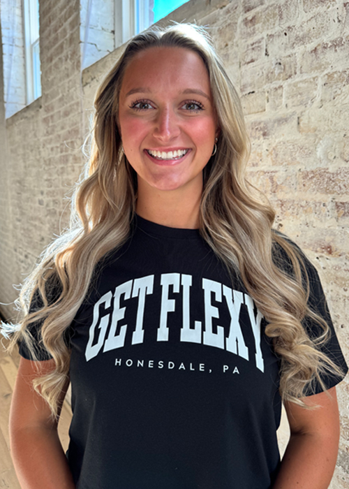 A woman with long wavy blonde hair smiles at the camera. Hannah Mascari, Client Relations Ambassador & Coach at Get Flexy, wears a black T-shirt that says GET FLEXY HONESDALE, PA, standing before a white brick wall with sunlight streaming in.