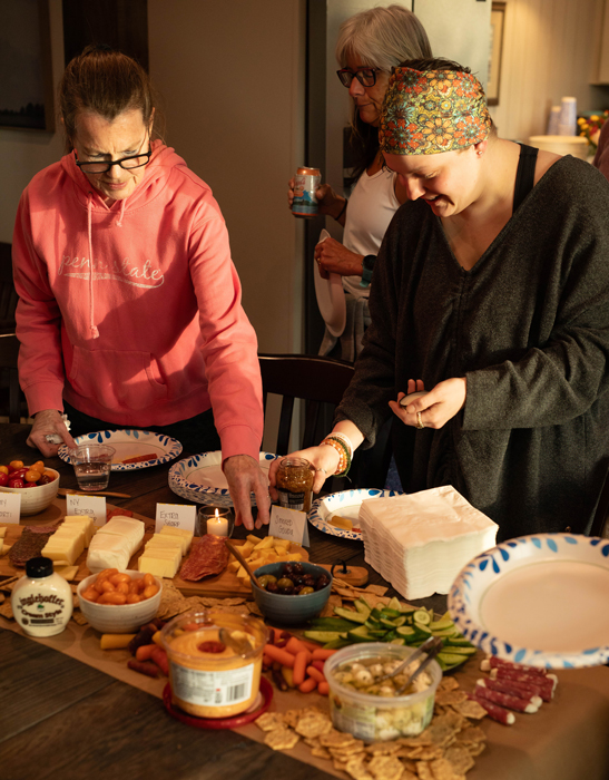 Two women prepare food at a table covered with cheeses, dips, crackers, and vegetables. Nutritionist at Get Flexy Riley Skelton’s expertise inspires the spread, while another woman in the sunlit room stands holding a drink.