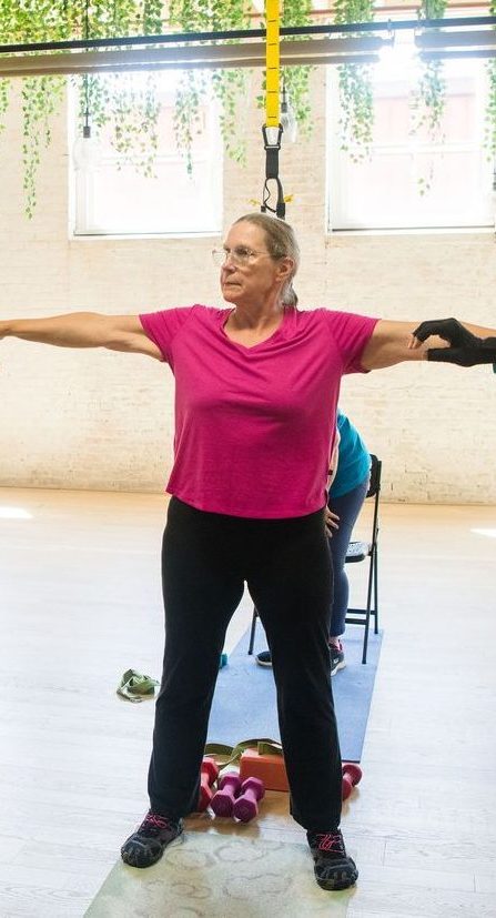 An older woman wearing a pink shirt and black pants stretches her arms out in a fitness studio, using suspension straps for support. Exercise equipment is on the floor nearby.