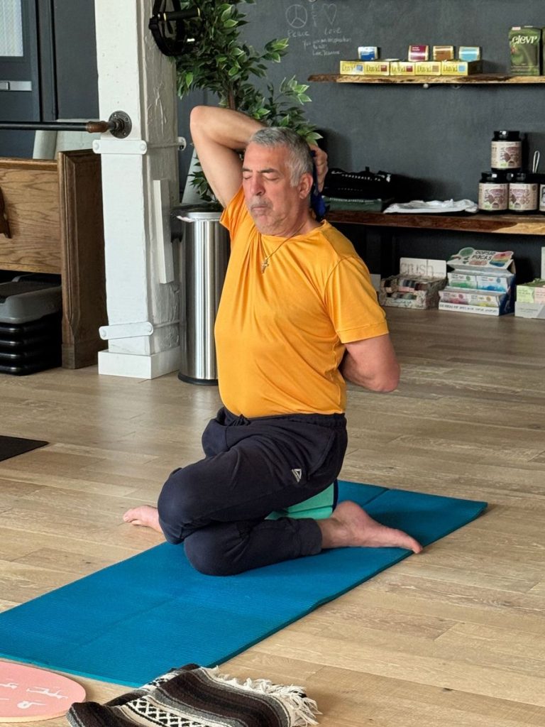 A man in an orange shirt and dark pants is practicing a seated yoga stretch indoors, holding a towel behind his back with both hands. He sits on a blue mat with one knee up, looking focused and calm.