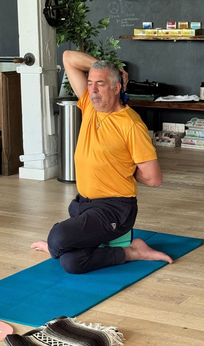 A man in an orange shirt and dark pants is practicing a seated yoga stretch indoors, holding a towel behind his back with both hands. He sits on a blue mat with one knee up, looking focused and calm.