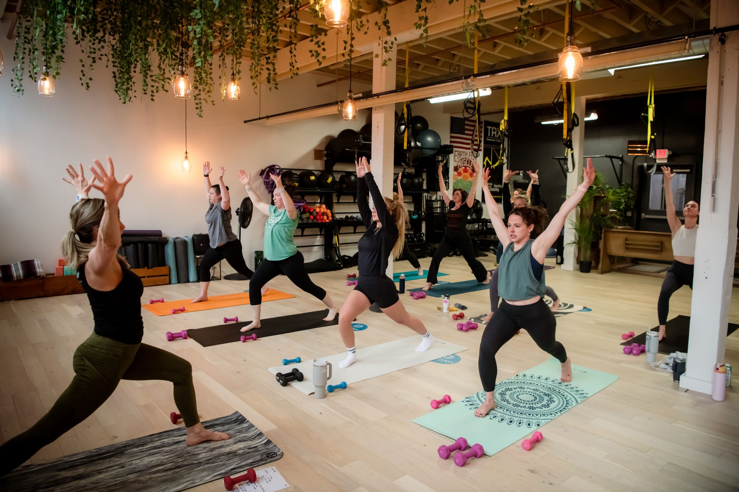 A group of people in workout clothes practice yoga poses on mats in a bright studio with wooden floors, hanging plants, and fitness equipment along the walls. Small dumbbells are placed beside their mats.