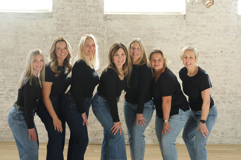 Seven women stand indoors in front of a white brick wall, all wearing black tops and blue jeans, smiling and posing closely together in bright natural light from windows above.