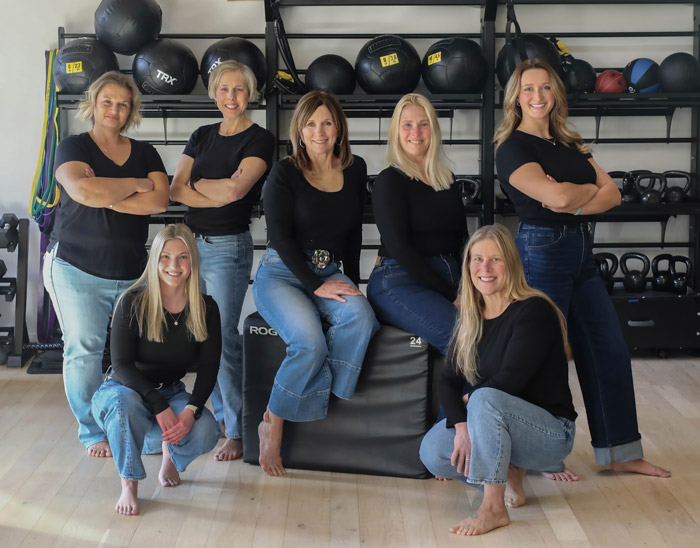 Seven women in black tops and blue jeans pose barefoot in a gym, surrounded by kettlebells, exercise balls, and resistance bands. Some are standing with arms crossed, while others are seated or kneeling, all smiling.