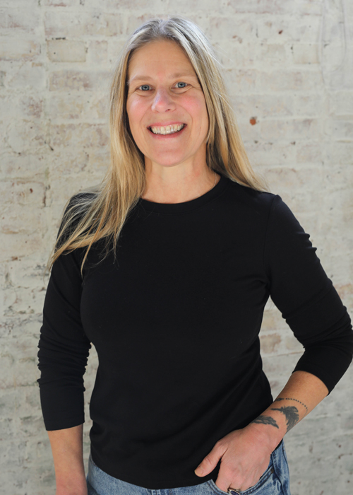 A smiling woman with long blonde hair wearing a black long-sleeve shirt and jeans stands in front of a light-colored brick wall, with one hand on her hip and a tattoo on her forearm.