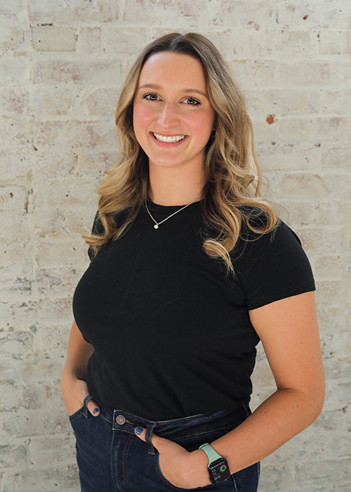 A woman with wavy blonde hair wearing a black t-shirt, dark jeans, a necklace, and a smartwatch stands smiling with her hands in her pockets in front of a light-colored brick wall.