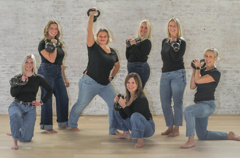 Seven women in black shirts and blue jeans pose barefoot indoors, each holding a kettlebell. They are smiling and standing against a white brick wall on a light wooden floor.
