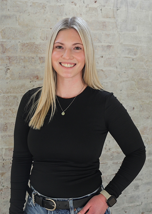 A woman with long blonde hair, wearing a black long-sleeve shirt, jeans, and a belt, stands smiling in front of a light-colored brick wall.